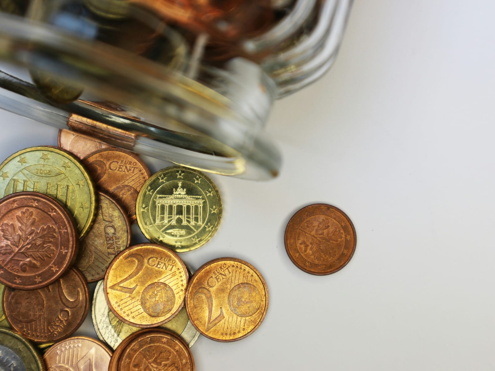 Euro coins spilling from a tipped glass jar onto a plain white surface, with one copper coin sitting apart from the pile
