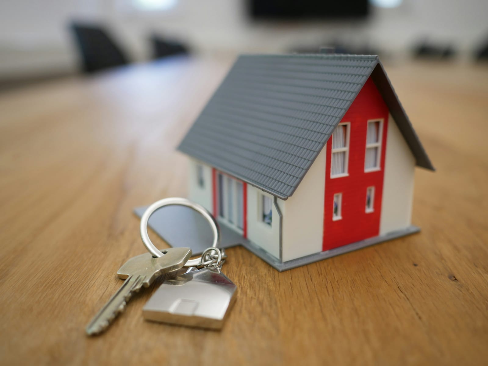 A small model house with a grey roof and red front beside a set of keys on a house-shaped keyring, resting on a wooden table